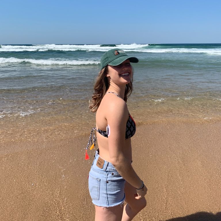 A photo of me (a white woman with dirty blond hair) standing on the beaching wearing a baseball hat, a bikini top, and denim shorts. I am standing to the side and looking over my shoulder at the camera, you can see my dexcom on my arm. The sky is a hazy light blue, the ocean is a deep teal with bright white waves, and the sand is beige.